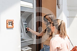 Two beautiful tourists female withdrawing the money from a bank card using ATM machine at the city center. Two travelers friend