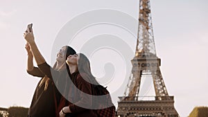 Two beautiful traveling female taking photos on smartphone near the Eiffel tower in Paris, France in sunny day.