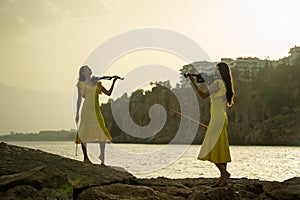 Two beautiful twin sisters violinists in yellow concert dresses are playing electric violins on sunset on rocks by the