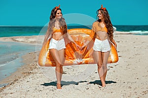 Two Beautiful twins girls in white shorts with orange Inflatable pool float mattress having fun on tropical beach.  Brunette