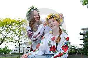 two beautiful Ukrainian girls weave wreaths on street embroidered shirts greenery nature sit near house Ukrainian