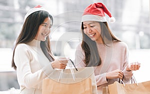 Two beautiful women wearing Santa hat and casual clothes, smiling with happiness, holding shopping paper bags on sales promotion