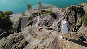 Two beautiful women in white dresses are photographed at the top of the mountain. One lady stands on the edge of cliff