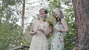 Two beautiful women with wild flowers standing at wooden stairs, talking, smiling. Girls looking at amazing nature. Slow