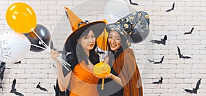 Two beautiful young asian woman in witch halloween costume holding pumpkin Jack O Lantern and balloon on white brick background