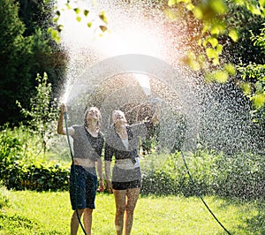 Two beautiful young cheerful woman pouring water from a sprinkler to themselfs and having fun in a green garden on a hot summer photo