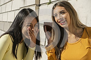 Two beautiful young Latina women giggling when listening to a funny audio message on their cell phone. Technology and