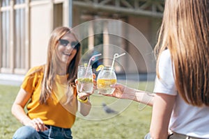 Two beautiful young woman in the summer sunny city drinking a cocktail from glass jars. Sitting on grass in park. Two laughing
