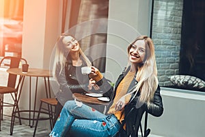 Two beautiful young women in fashion clothes having rest talking and drinking coffee in restaurant outdoor.