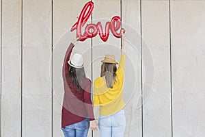 Two beautiful young women having fun outdoors with a red balloon with a love word shape. Casual clothing. They are wearing hats