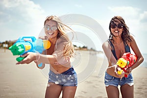 Two beautiful young women with sunglasses playing with water-gun on sandy beach