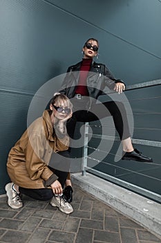 Two beautiful young women in trendy denim clothes with a leather jacket in sunglasses posing near a metal wall outdoors