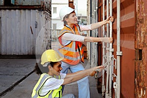 Two beautiful young worker engineer women wear safety vest and helmet, try to open shipping container door at logistic cargo yard