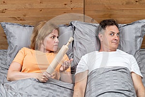 Two in bed, a woman holding a wooden rolling pin in her hand, a man scared. Relationship concept