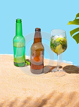 Two beer bottles and a cocktail glass with fresh mojito on the sandy beach with palm leaf shadow, and natural monstera on side.