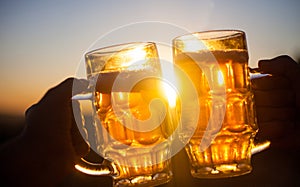 Two beer glasses in hands against blue sky and sun in the evening. Outdoor party, picnic concept