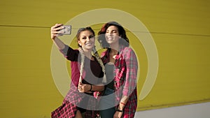Two best friends girls having fun and making selfie standing by the yellow wall. Two hipster girls taking selfie photos