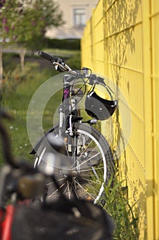 Two bicycles next to a yellow fence in the sunlight. Bikes with baskets and helmets in a green park with grass and trees in the
