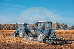 Two big blue tractor plowing a field and remove the remains of previously mown corn.