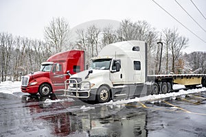 Two big rig long haul semi truck tractors with flat bed semi trailers standing side by side on the winter wet truck stop with snow