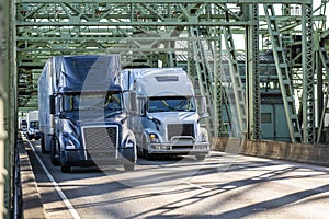 Two big rig semi trucks blue and white transporting cargo in dry van semi trailers running side by side on the truss arched bridge