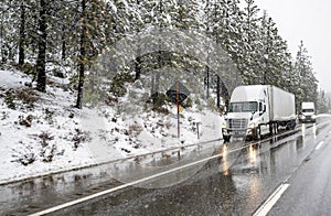 Two big rig semi trucks with semi trailers standing on the wet highway road shoulder waiting for the end of the winter snowing