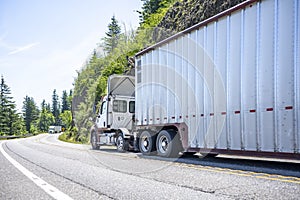 Two big rigs semi trucks with different semi trailers running towards each other on the winding mountain road with cliff and trees