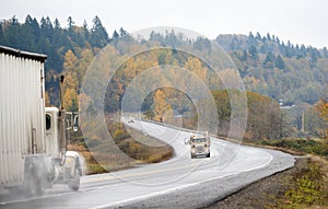 Two big rigs semi trucks move towards each other on winding road in the rain weather picking up cloud of rain dust from the road