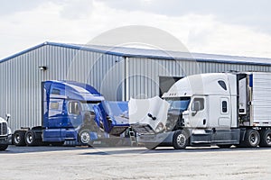 Two big rigs semi trucks with open hoods stand on the parking lot of the repair shop for engine maintenance and inspection and