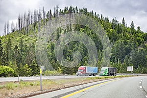 Two big rigs semi trucks transporting cargo in semi trailers driving side by side on the highway road along the forested mountain