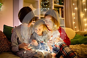 Two big sisters and their toddler brother playing with Chistmas lights in a cozy living room on Christmas eve. Kids spending time