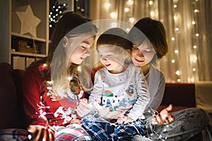 Two big sisters and their toddler brother playing with Christmas lights in a cozy living room on Christmas eve. Kids spending time
