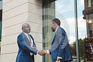 Two black african american businessmen in suits and glasses shake hands while meeting outdoors