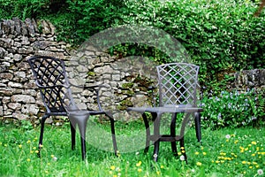 Two black chairs and black table is near stone wall on green  grass and dandelions in the garden in summer