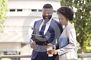 Two black coworkers checking financial reports outdoors, woman having coffee