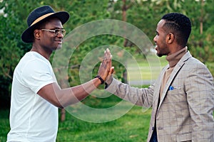 Two black men in stylish suits meeting in a summer park. African-Americans friends hispanic businessman embrace high