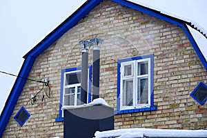 Two black metal chimney pipes on the roof under the snow at the front of a brick wall with windows