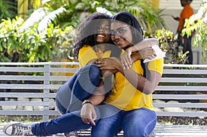 Two black women 20-25 years old together with curly hair happy in the park, sitting on a bench