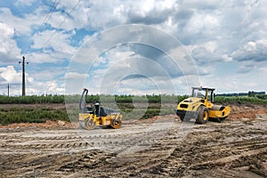 Two black and yellow road asphalt rollers stand on the ground, streaked with tracks from the tires of working machines