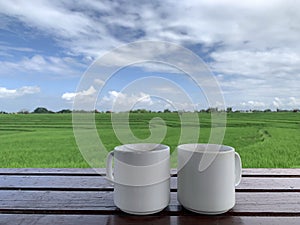 Two blank white mugs of tea or coffee on wooden table, with bright blue sky clouds over the fresh green field background.