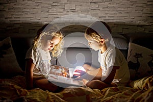 Two blond cute children, boy and girl, siblings, lying under the cover in bed, reading book together with small light