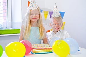 Two blonde caucasian kids boy and girl in birthday hats looking at camera and smiling at birthday party. Colorful