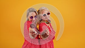 Two blonde excited middle-aged women wearing same red dotted dresses and sunglasses holding colorful lollipops. Yellow