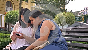 Two blubber caucasian girlfriends looking closely at phone and pressing on it chilling on bench outside