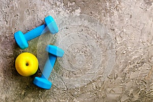 Two blue weights, dumbbells and yellow apple on concrete background.