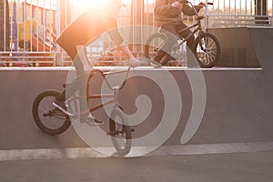 Two bmx cyclists riding a bike on a park skate on the background of the sunset