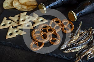 Two Bottles of beer with chips, salted crackers, bretzel and dried smelt fish on dark stone background