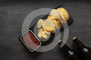 Two Bottles of beer with chips and tomato ketchup on dark stone background