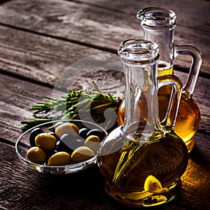 Two bottles of olive oil, olive in a bowl and herbs on a wooden table.