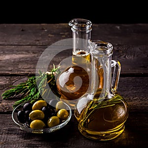 Two bottles of olive oil, olive in a bowl and herbs on a wooden table.